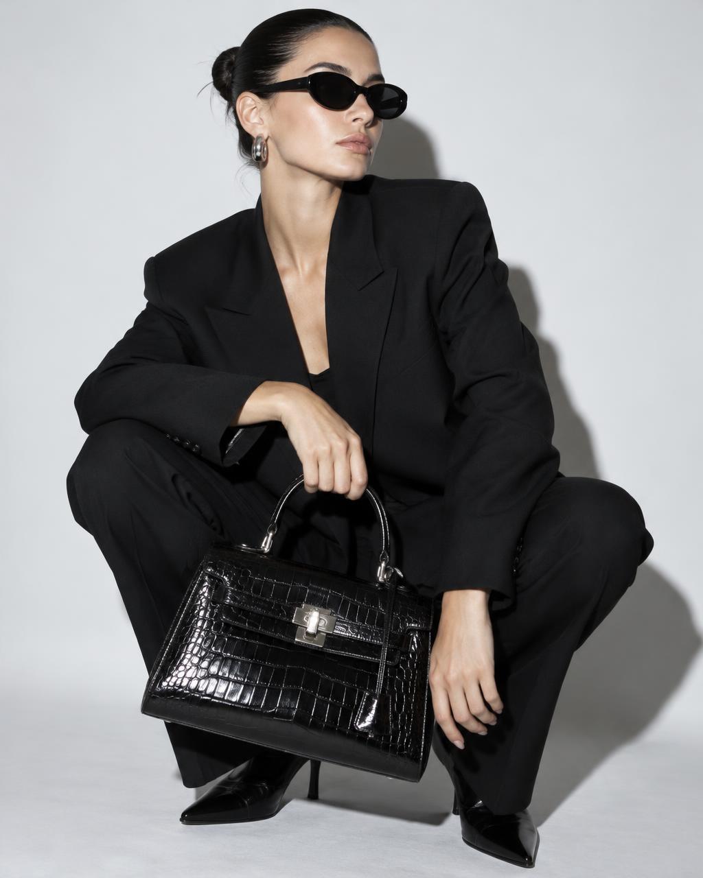 Model in black sunglasses and a black suit crouching with a glossy black croc-effect top-handle bag